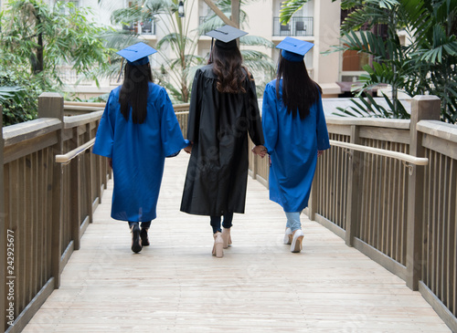 Three girl friends graduate walking on bridge wearing blue black graduation cap and gown holding hands indoors