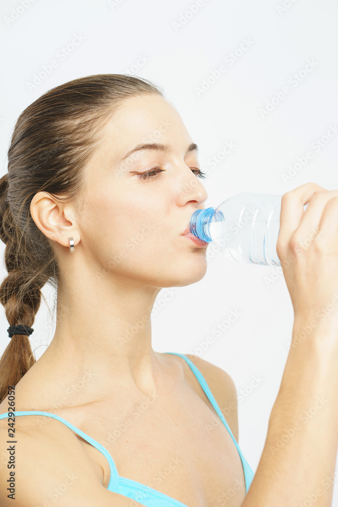 Attractive slim girl in sportswear drinks clear water from a plastic bottle during a workout on a white background. Attractive slim girl in sportswear drinks clear water from a plastic bottle during a workout on a white background.