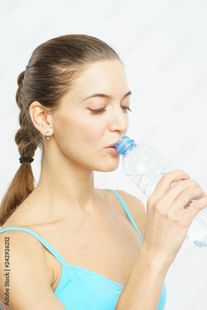 Attractive slim girl in sportswear drinks clear water from a plastic bottle during a workout on a white background. Attractive slim girl in sportswear drinks clear water from a plastic bottle during a workout on a white background.