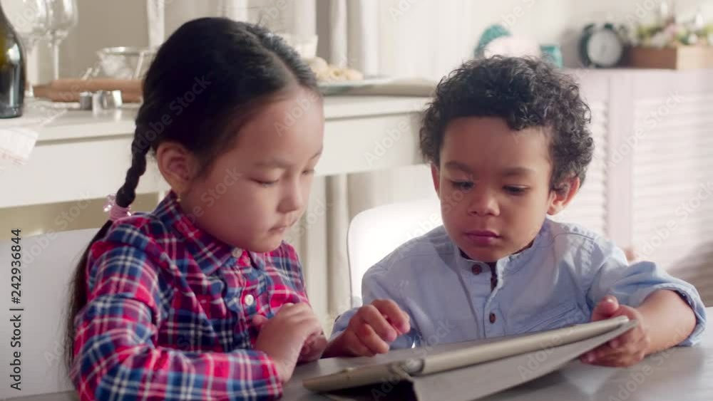 Little Asian girl and African American boy sitting together at kitchen table in home and surfing the Internet on digital tablet Little Asian girl and African American boy sitting together at kitchen table in home and surfing the Internet on digital tablet