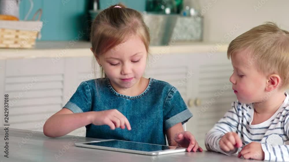 Cute little brother and sister sitting together at kitchen table in home and playing on digital tablet Cute little brother and sister sitting together at kitchen table in home and playing on digital tablet