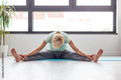 Mature woman in joga pose exercise in appartment at rug with window on background