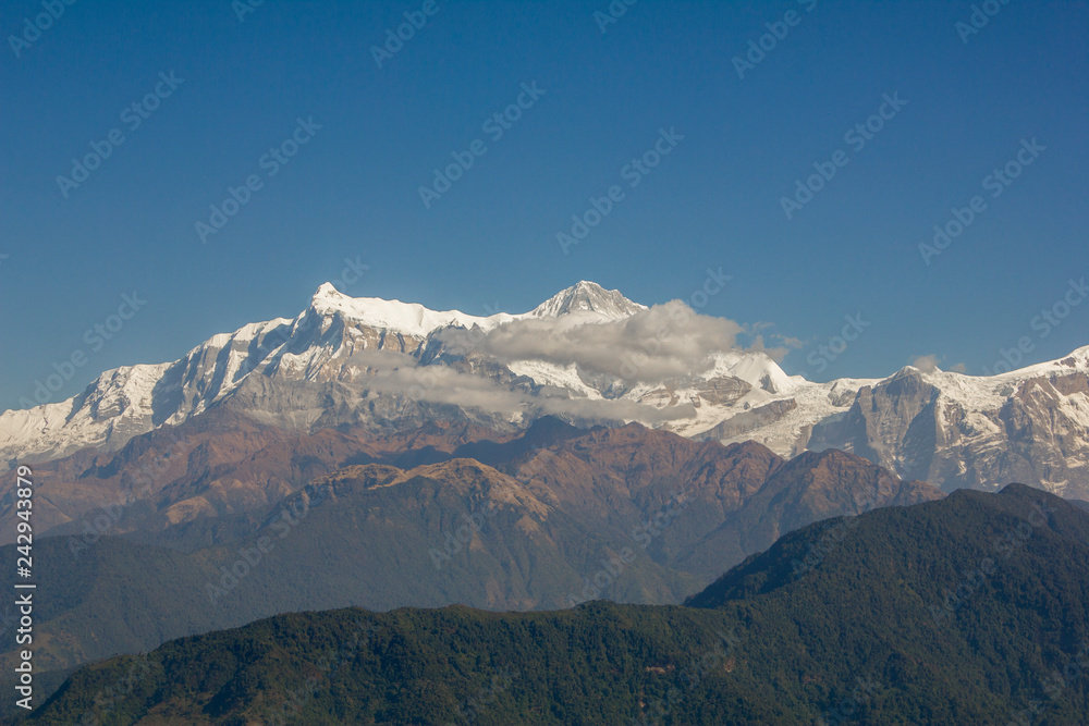 green slope of a mountain against the backdrop of clouds and the Annapurna snow ridge under a clear blue sky