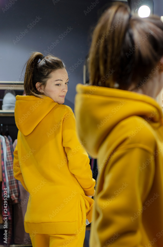 Young woman wearing bright yellow clothes (warm golf or sweater or tracksuit) standing near clothes and mirror rack indoors. Stylish wardrobe Young woman wearing bright yellow clothes (warm golf or sweater or tracksuit) standing near clothes and mirror rack indoors. Stylish wardrobe