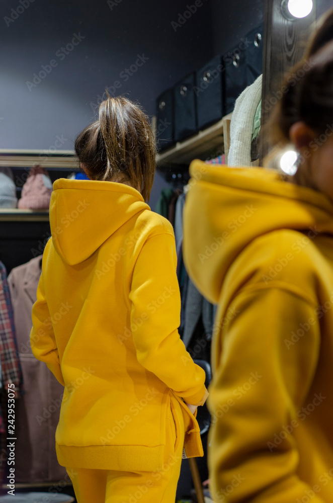 Young woman wearing bright yellow clothes (warm golf or sweater or tracksuit) standing near clothes and mirror rack indoors. Stylish wardrobe Young woman wearing bright yellow clothes (warm golf or sweater or tracksuit) standing near clothes and mirror rack indoors. Stylish wardrobe