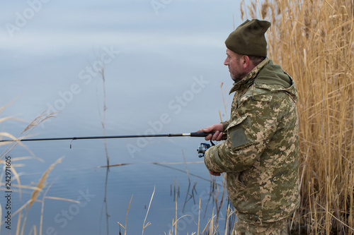 Fishing in the reeds for spinning on quiet water. A man in camouflage clothes in cool weather is fishing on the river bank.