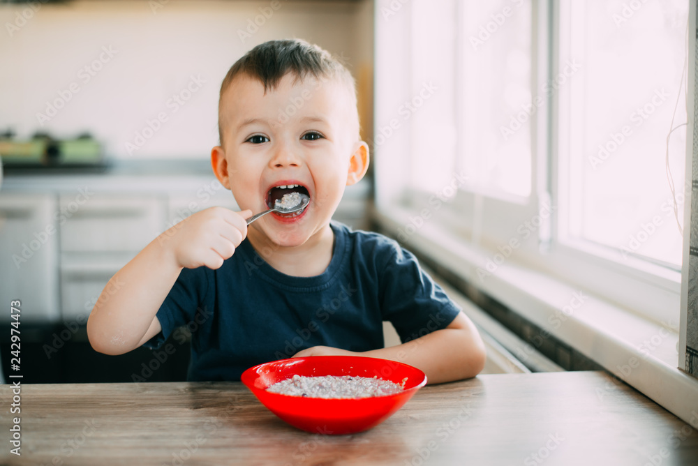 A child in the kitchen eating their own oatmeal with a red plate A child in the kitchen eating their own oatmeal with a red plate