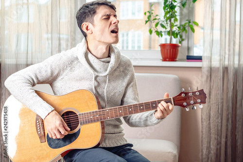 Young attractive male musician sitting on a chair playing acoustic guitar in room. Concept of music as a hobby