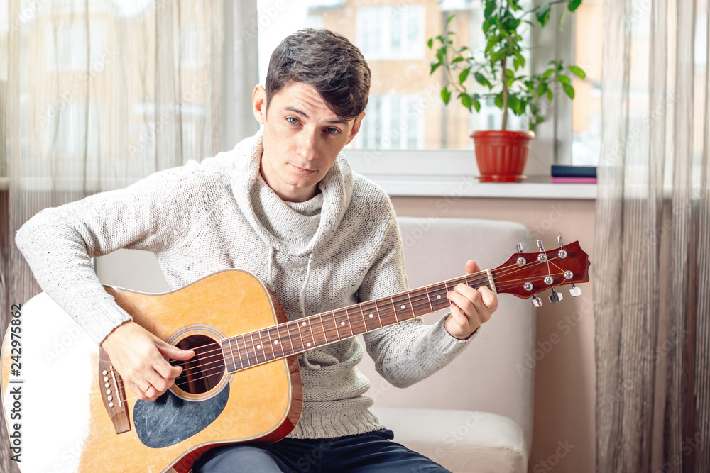 Obraz premium Young attractive male musician sitting on a chair playing acoustic guitar. Concept of music as a hobby