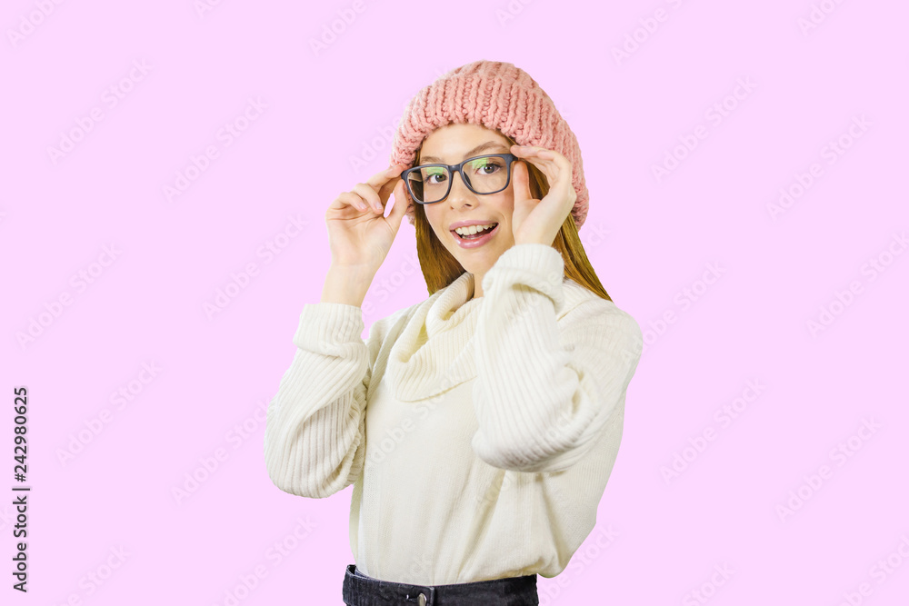Portrait of a young red-haired girl in a knitted pink hat who is holding glasses with both hands smiling looking at the camera against an isolated background Portrait of a young red-haired girl in a knitted pink hat who is holding glasses with both hands smiling looking at the camera against an isolated background
