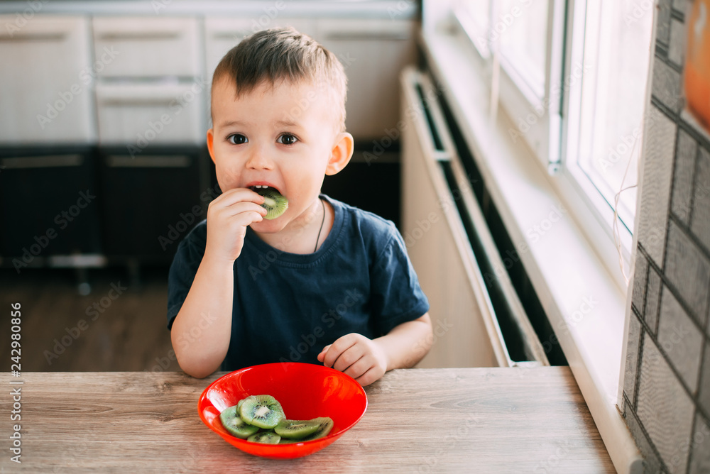 The boy at the table eating kiwi sliced into slices in a red plate The boy at the table eating kiwi sliced into slices in a red plate