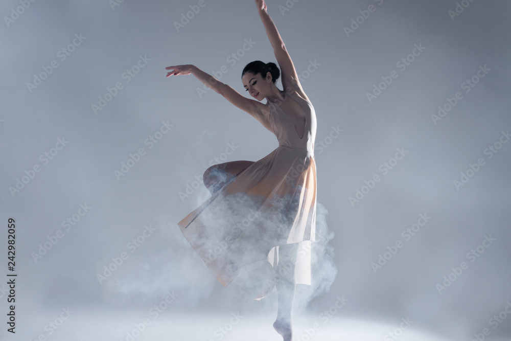 Fototapeta premium Elegant woman female girl ballerina dancer in beige dress dancing, making performance and dance element ballet in fog dust smoke fume on isolated grey background scene. Dancing in cloud concept