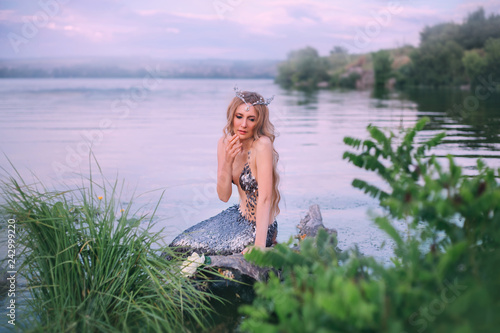 dreamy blonde nymph with a sea diadem sits alone in the river and looks down in surprise, raised her hand to her mouth, a dreamy lady with a silver tail against the blue sky and bright green grass
