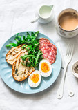 Delicious breakfast or snack - salami sausage, boiled egg, arugula, grilled bread and coffee on a light background, top view