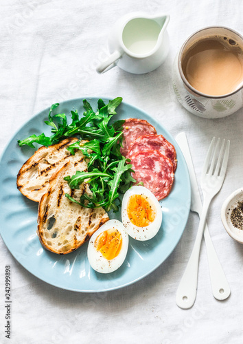 Delicious breakfast or snack - salami sausage, boiled egg, arugula, grilled bread and coffee on a light background, top view