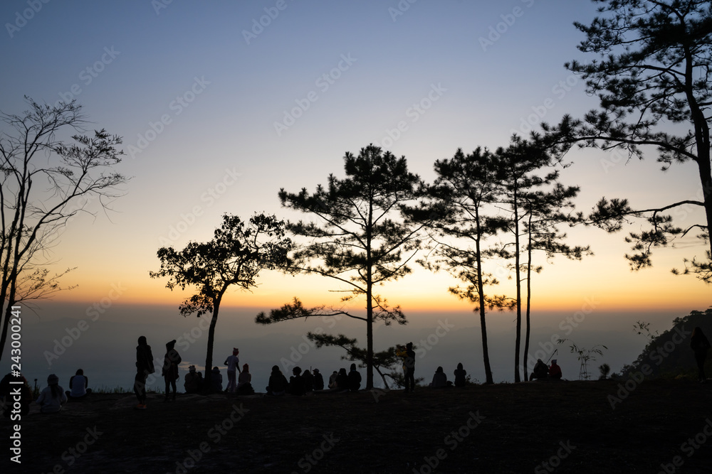 Image of sunrise on orange and yellow horizon with people's silhouette surrounded by pine trees ( Phu kradueng Thailand )