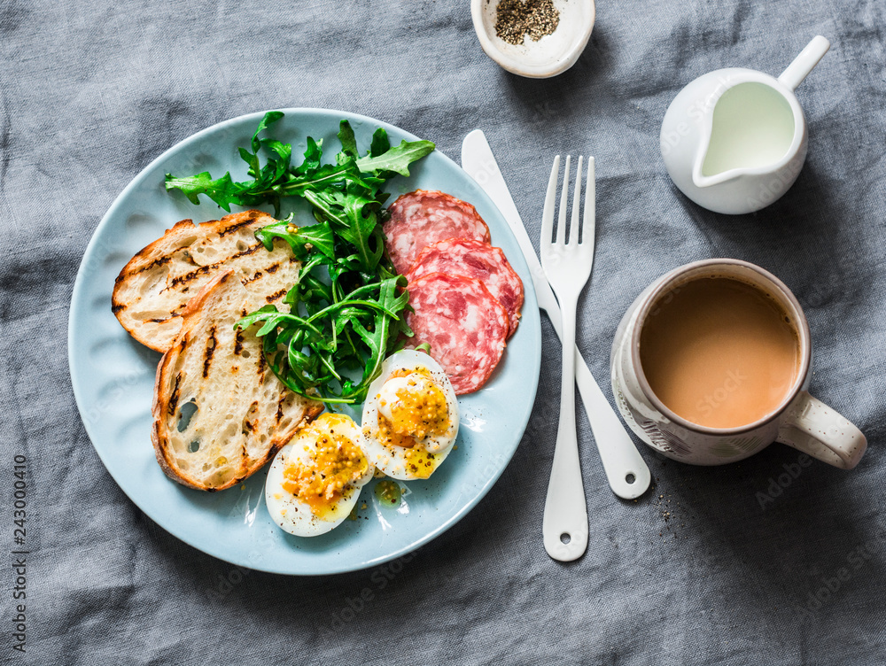 Delicious breakfast or snack - salami sausage, boiled egg, arugula, grilled bread and coffee on a gray background, top view