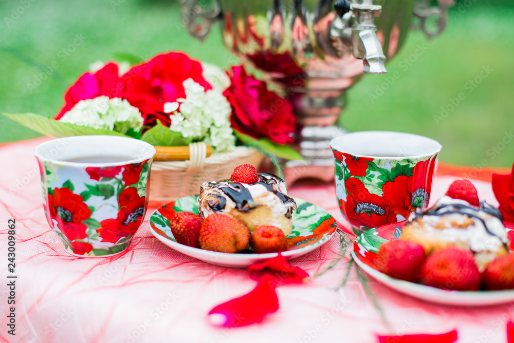 Samovar with a cup of morning tea, cinnamon buns and strawberry on the table on nature background Samovar with a cup of morning tea, cinnamon buns and strawberry on the table on nature background