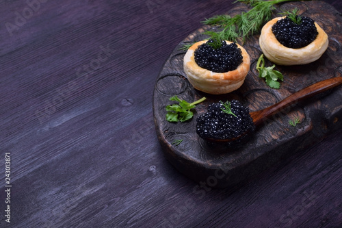 Black caviar in a wooden spoon and puff pastry tartlets on a dark board