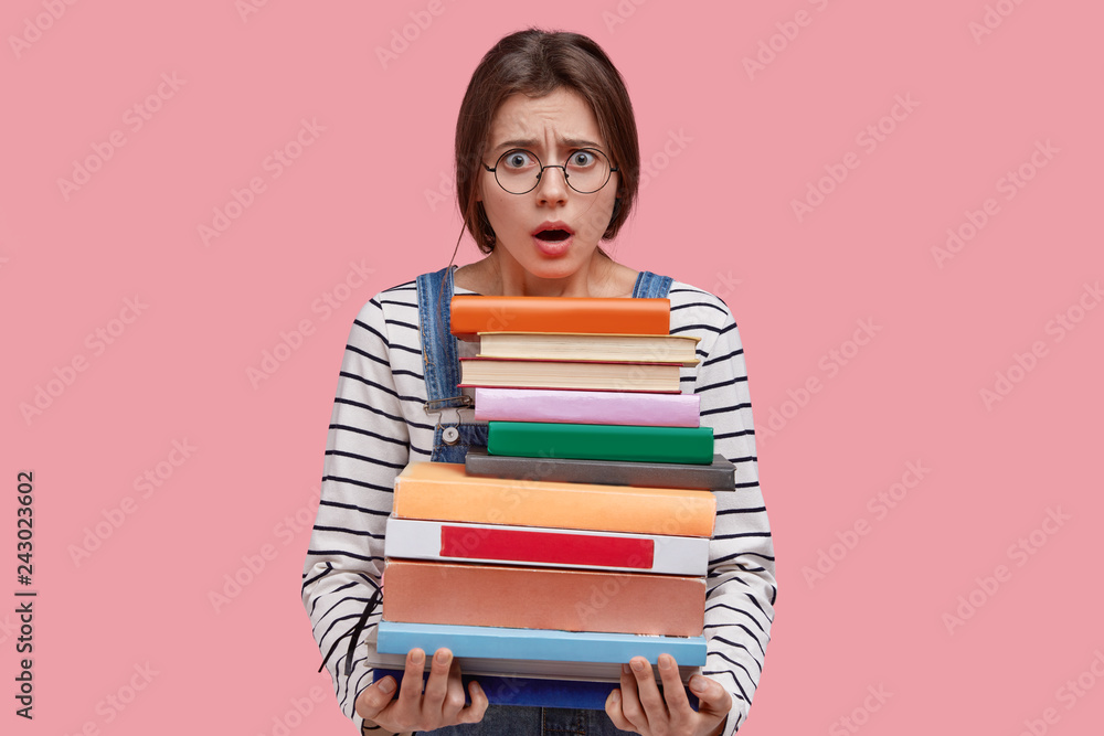 Photo of indignant young lady keeps jaw dropped from negative emotions, wears round spectacles, holds heavy pile of books, returns from library, prepares for final examination or test at college Photo of indignant young lady keeps jaw dropped from negative emotions, wears round spectacles, holds heavy pile of books, returns from library, prepares for final examination or test at college