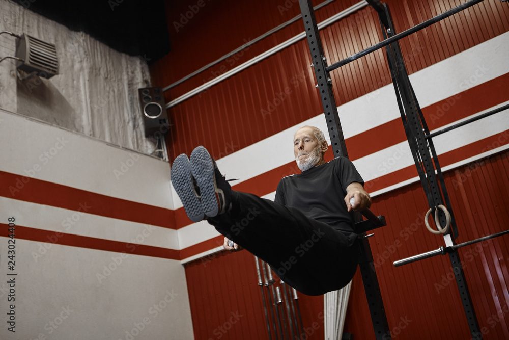 Low angle view of attractive elderly European male pensioner having focused look while doing vertical legs raise on horizontal bar at fitness center. Senior man exercising indoors with equipment