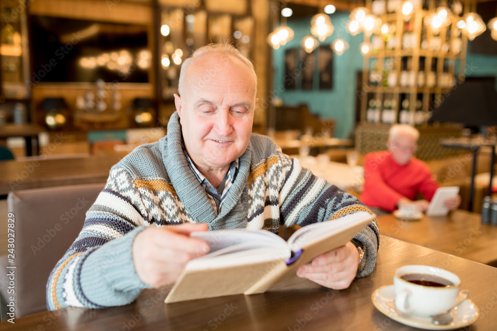 Content curious chubby mature man in casual sweater sitting at wooden table with coffee cup and reading interesting book in cafe Content curious chubby mature man in casual sweater sitting at wooden table with coffee cup and reading interesting book in cafe