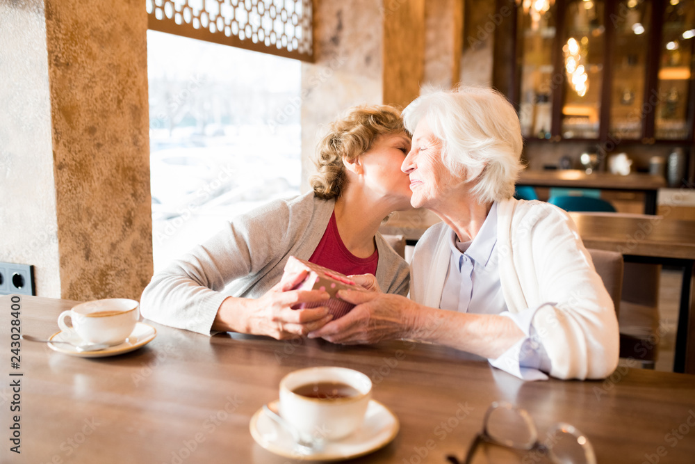 Content mature lady in cardigan sitting at table and kissing friend while giving gift to her in modern cafe Content mature lady in cardigan sitting at table and kissing friend while giving gift to her in modern cafe