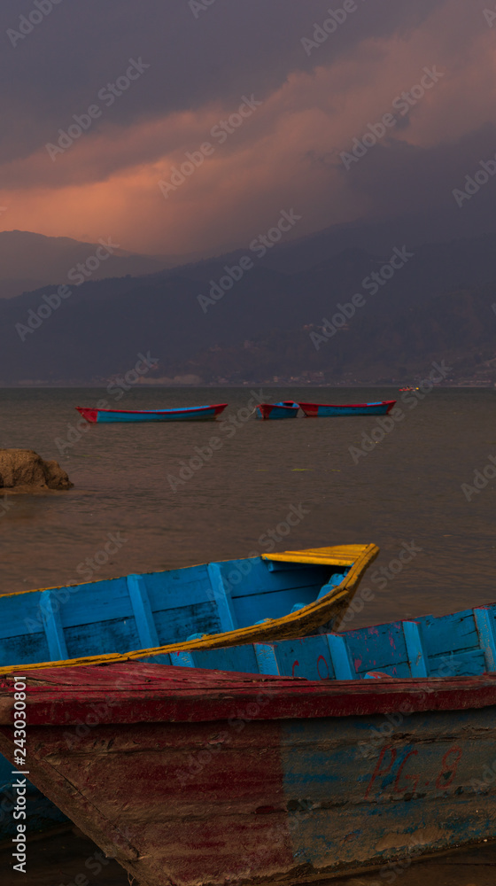 Boats in Pokhara lake in Nepal during sunset in a rainstorm. Sun shining through the clouds above the mountains in the background. 