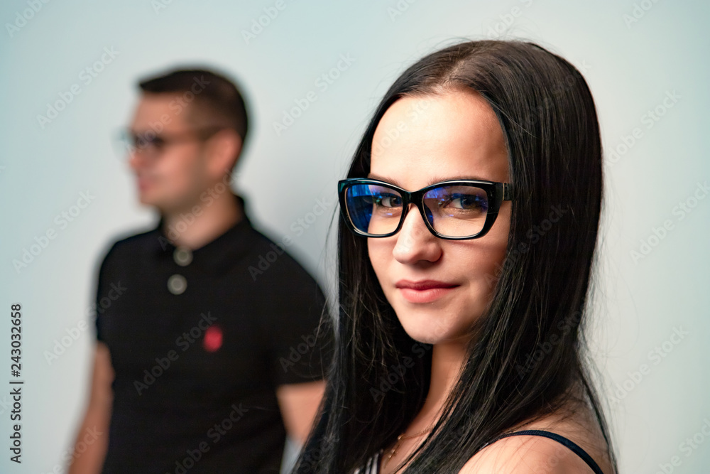Attractive female brunette with modern eyeglasses in black frame smiling in front of her boyfriend background. Pretty young woman wearing trendy glasses in studio. Close-up. Attractive female brunette with modern eyeglasses in black frame smiling in front of her boyfriend background. Pretty young woman wearing trendy glasses in studio. Close-up.
