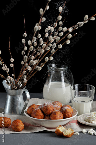 Fried in butter balls of cottage cheese. In the frame is a plate with cottage cheese and milk. Festive table setting supplemented willow twigs. Dark background. Vertical frame orientation.