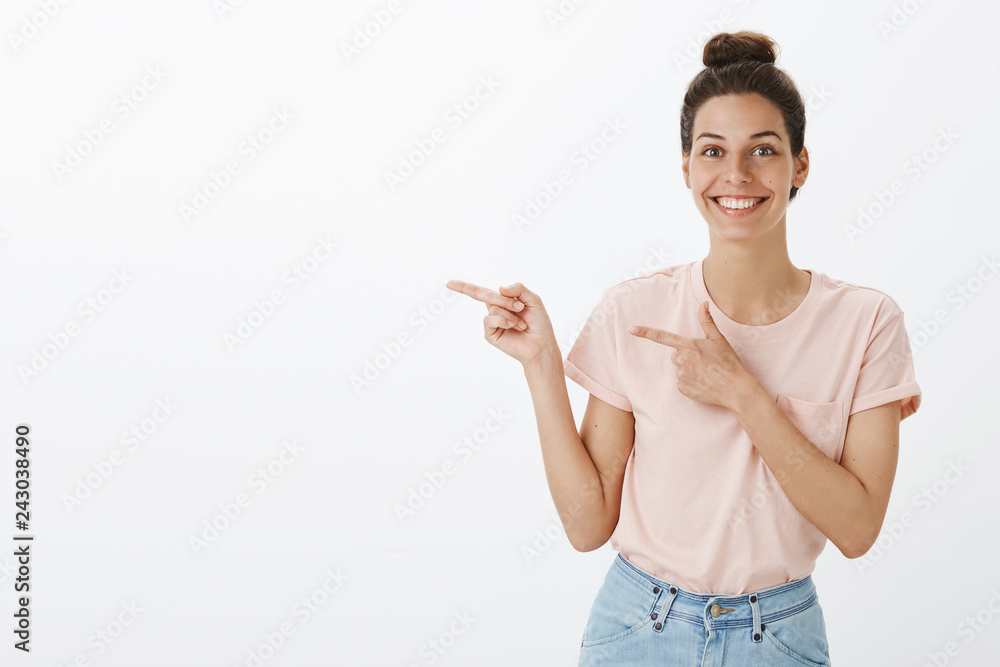 Waist-up shot of good-looking happy young charismatic european female in pink t-shirt and jeans with hairbun pointing left with index fingers and smiling impressed showing awesome copy space Waist-up shot of good-looking happy young charismatic european female in pink t-shirt and jeans with hairbun pointing left with index fingers and smiling impressed showing awesome copy space