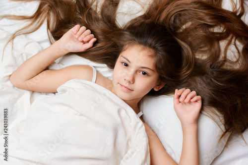 Playful pretty girl with a long natural dark hair lies on bed covered with white soft blanket. Adorable little child spread her beautiful hair on the confortable pillow on bed