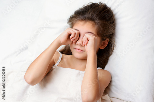 Sleepy pretty little child rubs her eyes in the morning. Lovely small child with long dark natural hair lies on comfortable bed rubbing eyes and yawning