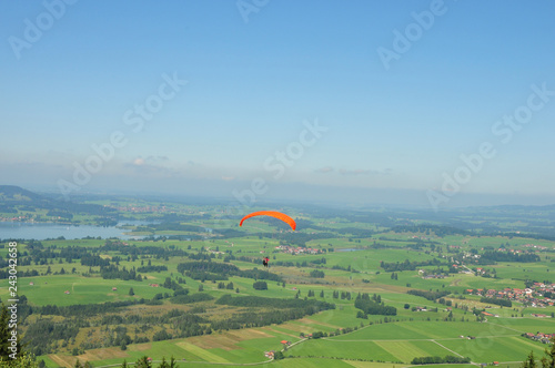 Paraglider with an orange umbrella gliding over a beautiful valley with blue sky