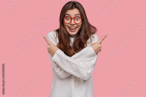 Joyful pretty woman points at different corners upwards, has happy facial expression, wears spectacles and white jumper, isolated over pink background. People, choice and hesitation concept.
