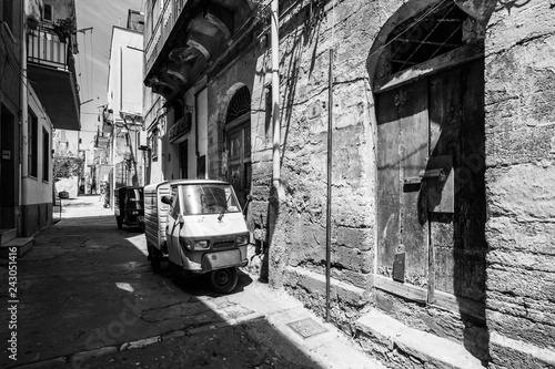 Fototapeta Naklejka Na Ścianę i Meble -  Black and white image of Sciacca old town with a Ape three-wheeled vehicle near old buildings. Sciacca, Sicily, Italy, May 2018