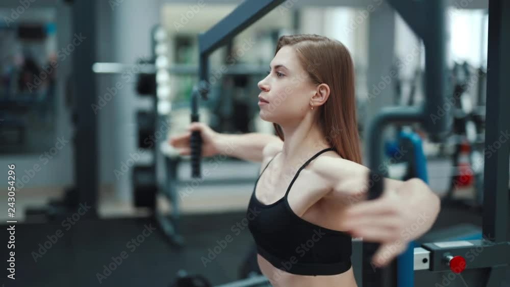 A beautiful girl with her strong arms performs an exercise on a simulator to strengthen the pectoral muscles in a fitness club. Close-up. Blurred Background A beautiful girl with her strong arms performs an exercise on a simulator to strengthen the pectoral muscles in a fitness club. Close-up. Blurred Background