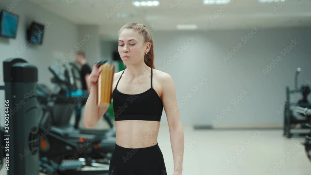 A slender woman in a black sports top and with her hair stuck in the tail is drinking water from a shaker and walking through the gym. Blurred Background A slender woman in a black sports top and with her hair stuck in the tail is drinking water from a shaker and walking through the gym. Blurred Background