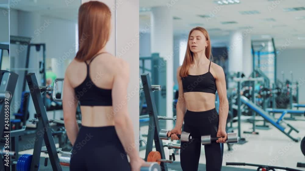 A beautiful woman in black leggings and top is doing an exercise for biceps using dumbbells opposite mirror in the gym. Blurred Background A beautiful woman in black leggings and top is doing an exercise for biceps using dumbbells opposite mirror in the gym. Blurred Background