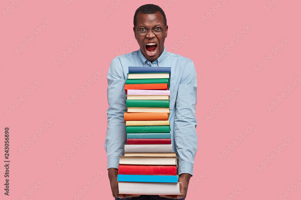 Photo of outraged black man yells in annoyance, reads textbooks, irritated with much tasks, dressed in formal clothes, expresses negative emotions, isolated over pink background. I hate studying Photo of outraged black man yells in annoyance, reads textbooks, irritated with much tasks, dressed in formal clothes, expresses negative emotions, isolated over pink background. I hate studying