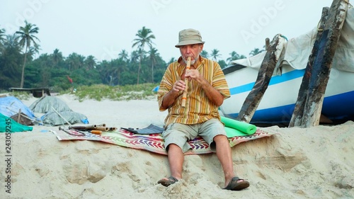 Senior man playing bamboo flute on the beach next to fishing boat