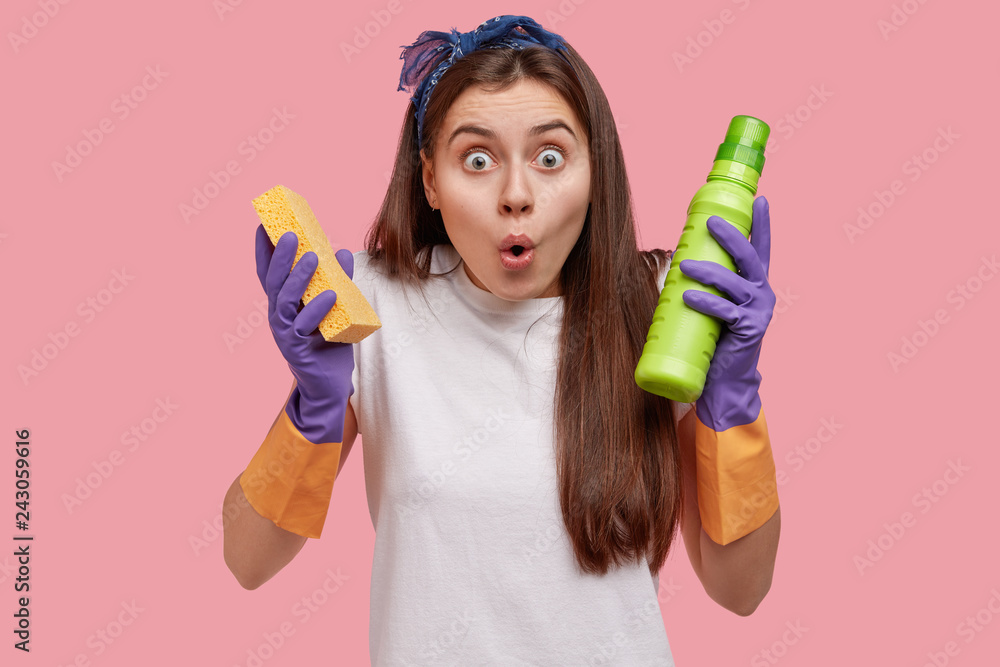 Shocked attractive young female from cleaning service, holds sponge and washing detergent, keeps jaw dropped, wears white t shirt, models over pink background. Cleaning and disinfect concept. Shocked attractive young female from cleaning service, holds sponge and washing detergent, keeps jaw dropped, wears white t shirt, models over pink background. Cleaning and disinfect concept.