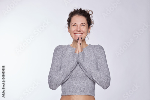 Cute blue eyed American woman keeps hands in praying gesture, has broad smile, looks up, preparing for important event, dressed in cashmere top, poses against white background. Believe in better. photo