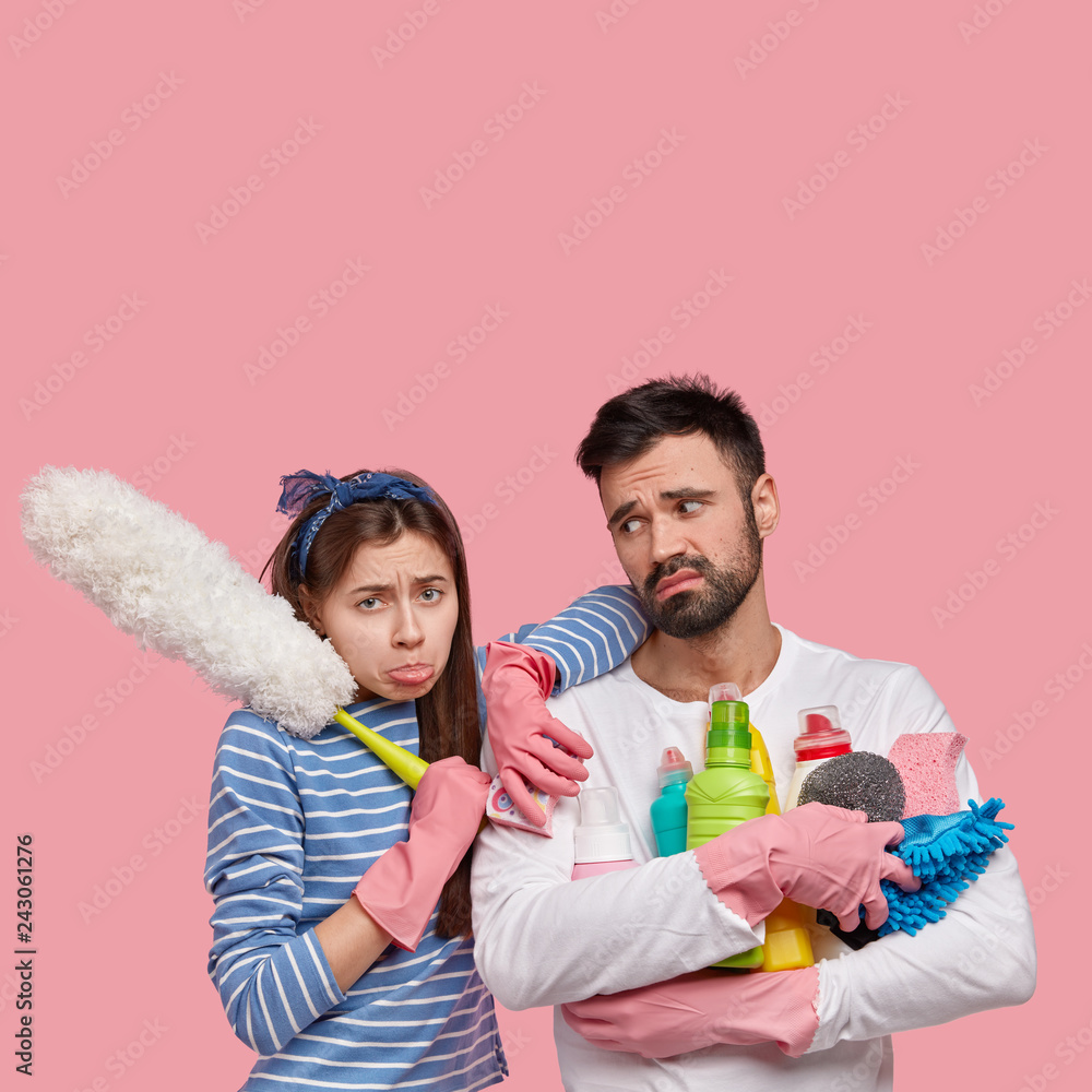Image of dejected wife and husband clean at home, do spring cleaning, feel fatigue, stand closely to each other, hold detergents, sponge and brush, isolated over pink wall with blank space above