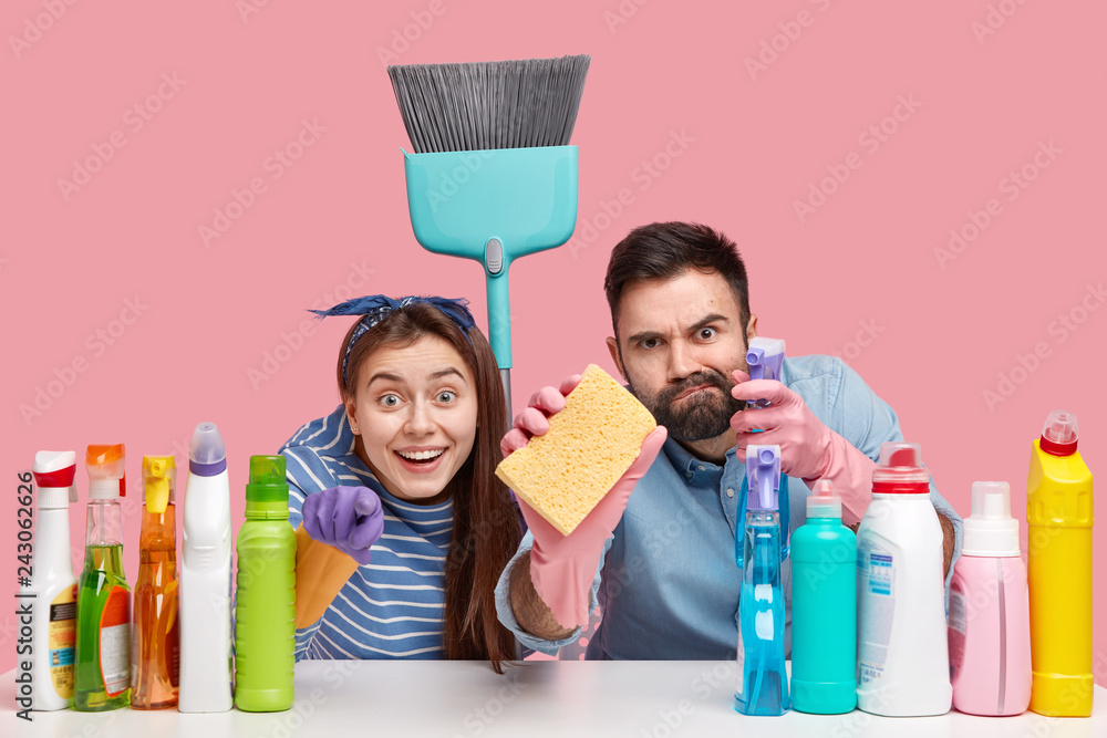 Positive two people look scrupulously at camera, hold broom and sponge, sit at desktop with cleaning supplies, notice object for cleaning, isolated against pink background. Tidying up concept