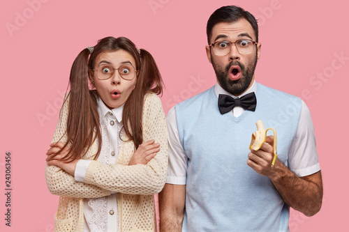 Photo of surprised female and male wonks stare with disbelief, eat delicious banana, dressed in old fashionable clothes, being puzzled by terrible news, isolated over pink background. Reaction concept photo