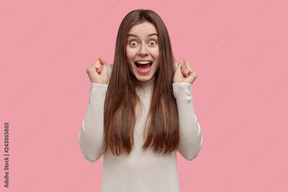 Positive European dark haired woman yells yes with success, clenches fists in triumph, celebrates victory, dressed in casual clothes, stands against pink background. People and body language concept Positive European dark haired woman yells yes with success, clenches fists in triumph, celebrates victory, dressed in casual clothes, stands against pink background. People and body language concept
