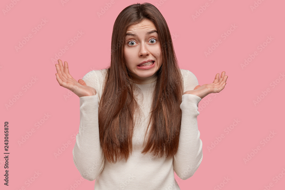 People, apathy and attitude concept. Questioned beautiful young woman shruggs shoulders with hesitation, feels doubt, has long dark hair, wears casual white jumper, isolated against pink wall People, apathy and attitude concept. Questioned beautiful young woman shruggs shoulders with hesitation, feels doubt, has long dark hair, wears casual white jumper, isolated against pink wall