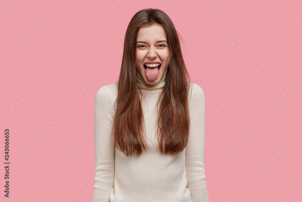 Comic young female model sticks out tongue, makes happy grimace, dressed in white clothes, has long hair, expresses positive emotions, stands against pink background, foolishes with friends.