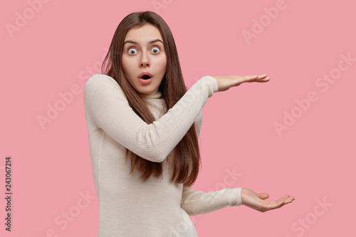 Stunned scared lady with bugged eyes shapes square tall huge object with both hands, gestures against copy space, dressed in white clothes, has long hair, isolated over pink studio background